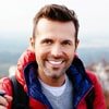 Smiling man in a red jacket outdoors with a blurred landscape in the background