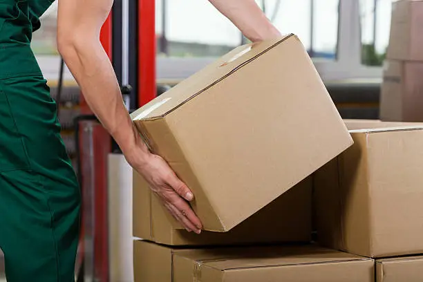 istockphoto-499617919-612x612-1 – ZAZALAND COUNTER 🚬 Worker in a green uniform lifting a brown cardboard box onto a stack in a warehouse setting