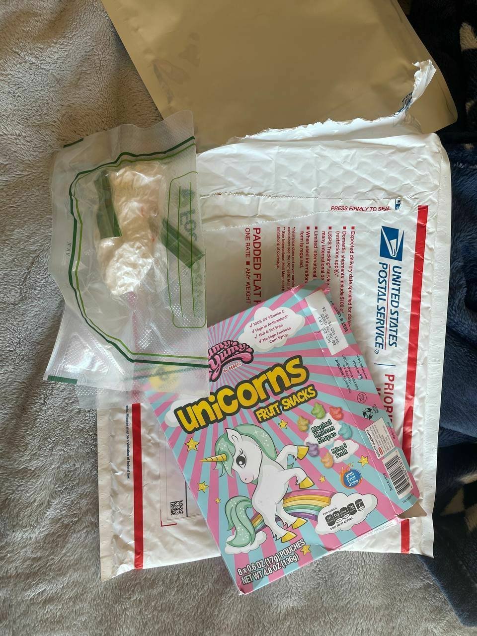 photo_2025-02-26_20-09-01 2 – ZAZALAND COUNTER 🚬 White USPS Priority Mail mailing envelope with a pink and teal aposUnicorns Fruit Snacksapos box resting on top packing materials visible nearby clear bag and brown paper